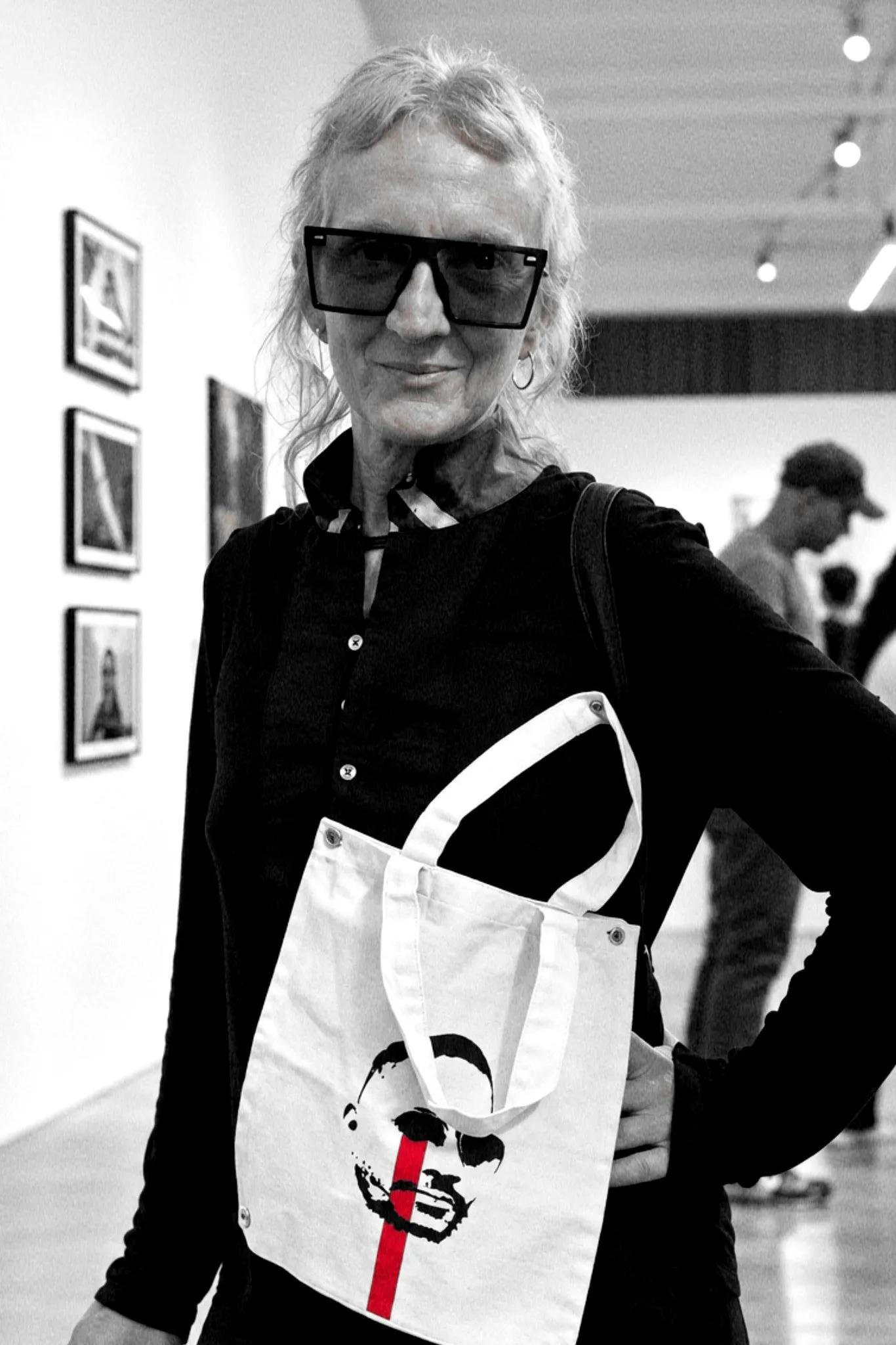 Retrato de Sandra Ceballos en una galería, con gafas de sol y una bolsa de tela con una cara y una raya roja.
